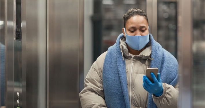 Señora abrigada dentro del ascensor con mascarilla y mirando el móvil