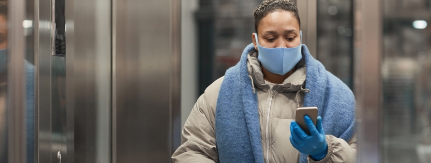Señora abrigada dentro del ascensor con mascarilla y mirando el móvil